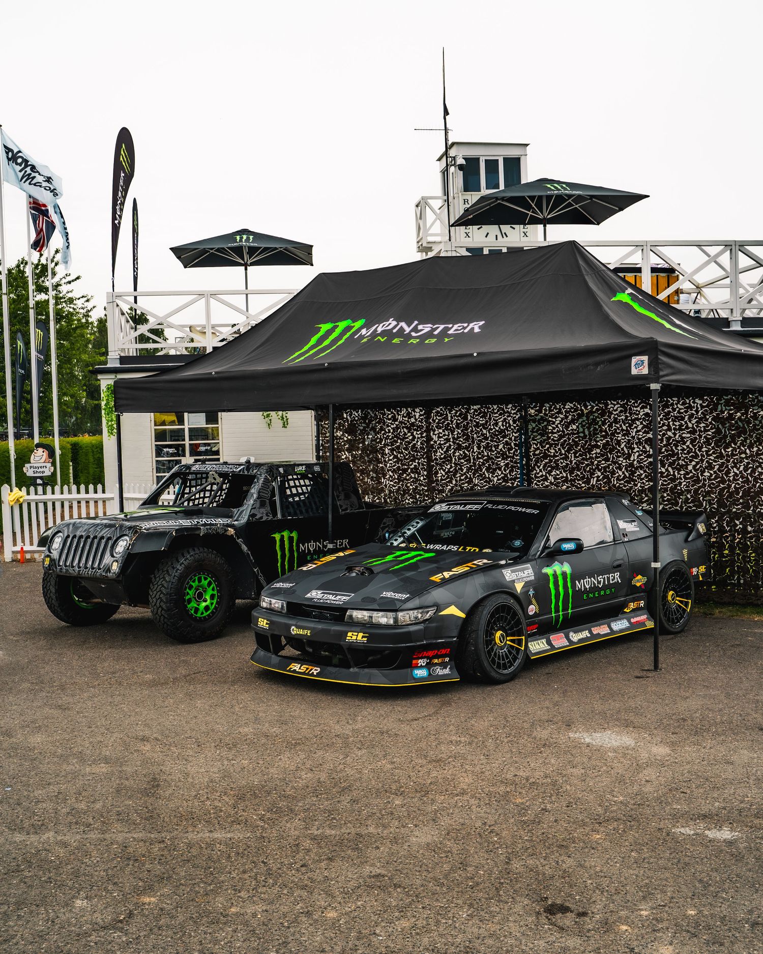 Black Monster Energy branded racing cars displayed under promotional tents at an outdoor event.