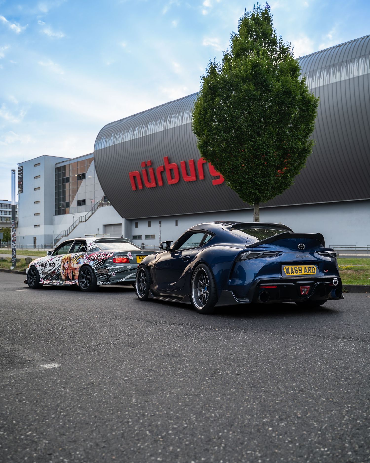 Row of sports cars parked outside a Nürburgring facility on a cloudy day.
