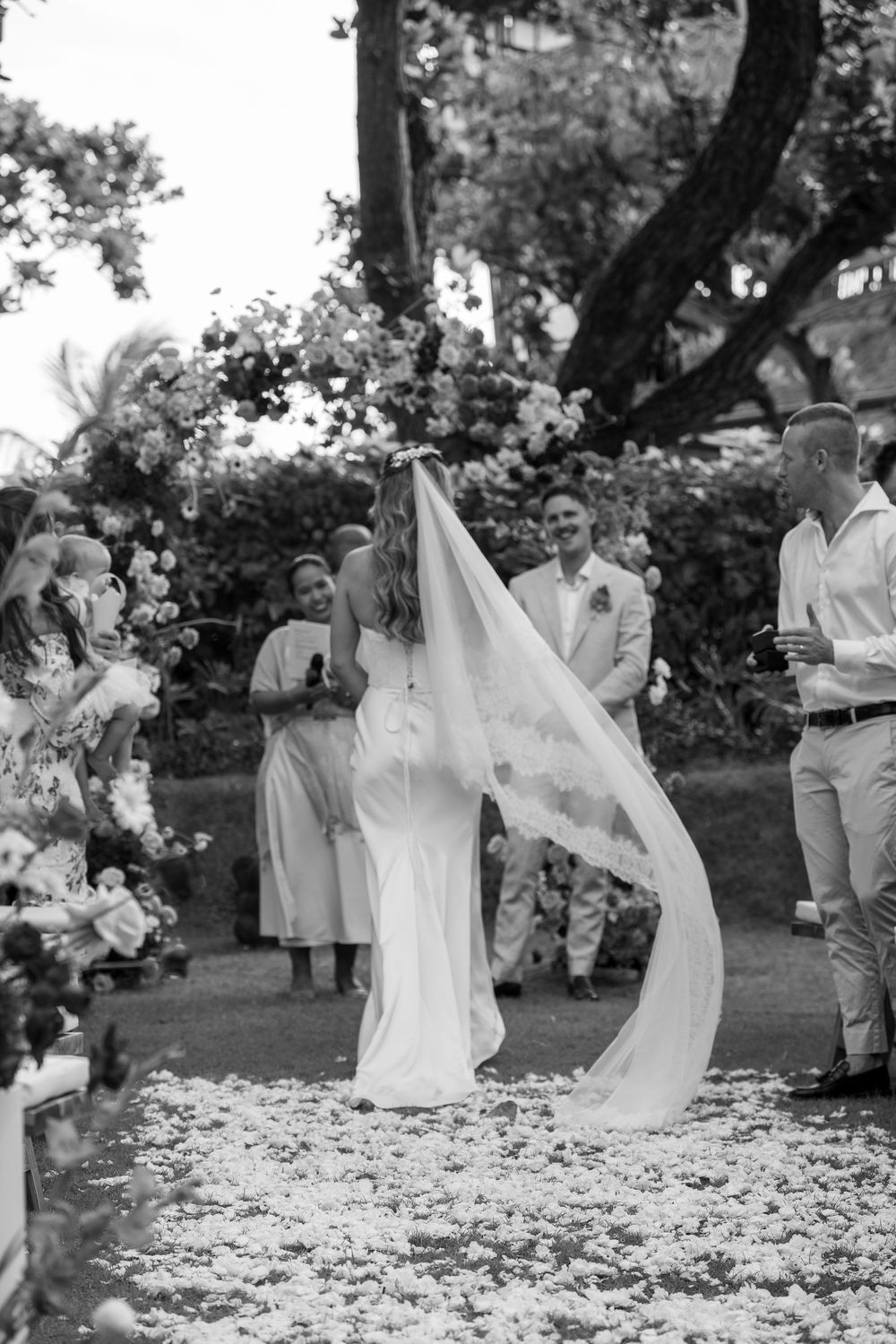 A black and white photos showing a bride walking through scattered flower petals in a garden wedding.