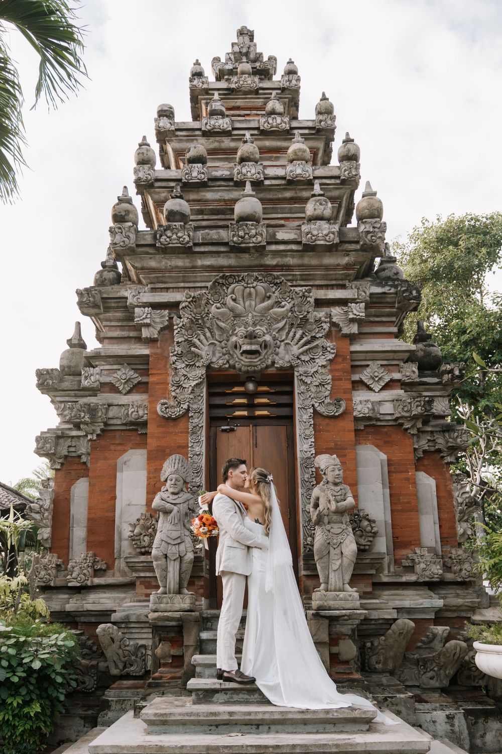 A couple shares a romantic moment in front of an ornate Balinese temple with traditional stone architecture and carvings.