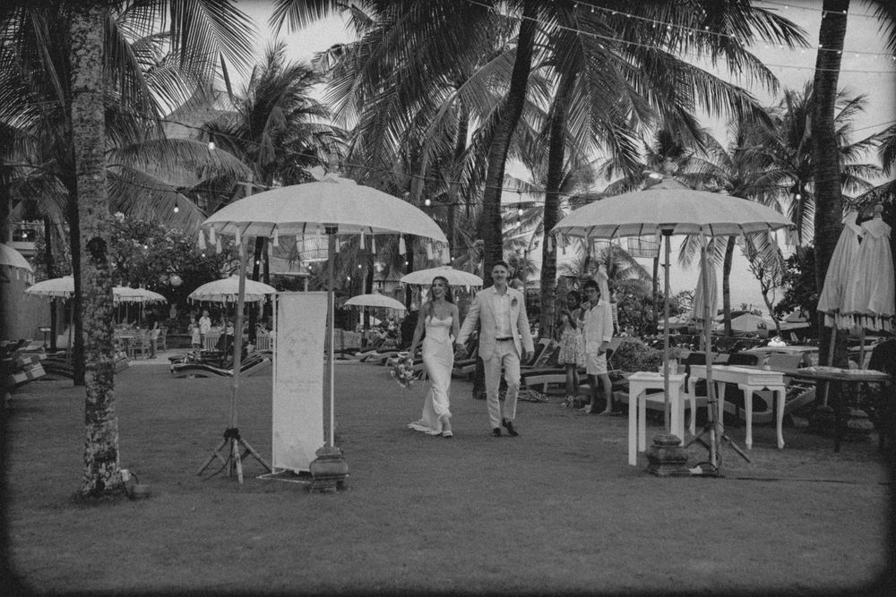 Vintage black and white photo showing a married couple enjoying a tropical resort setting with palm trees and umbrellas.