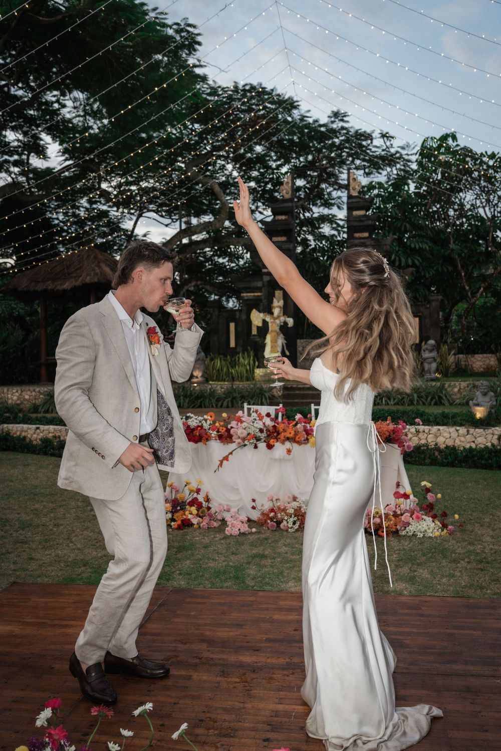 A newly married couple dancing joyfully together on a wooden deck with floral decorations in the background at an outdoor wedding