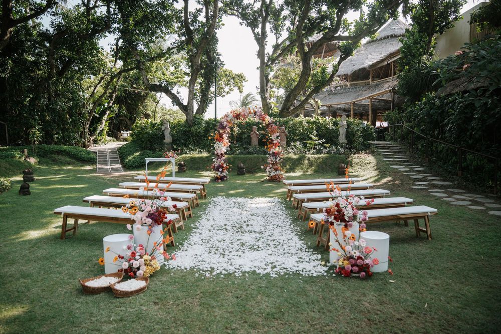 Outdoor garden wedding ceremony setup with wooden benches and floral arrangements under tall trees in Bali