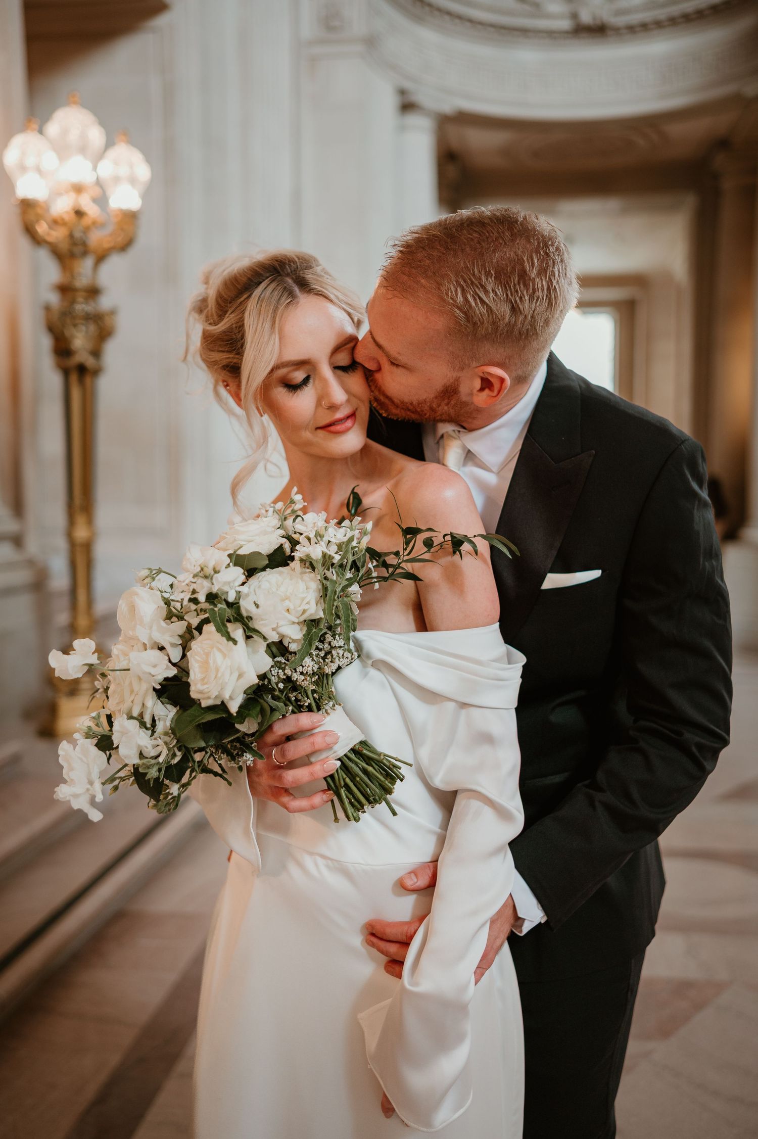 Romantic wedding couple embraces with white floral bouquet in elegant indoor setting.