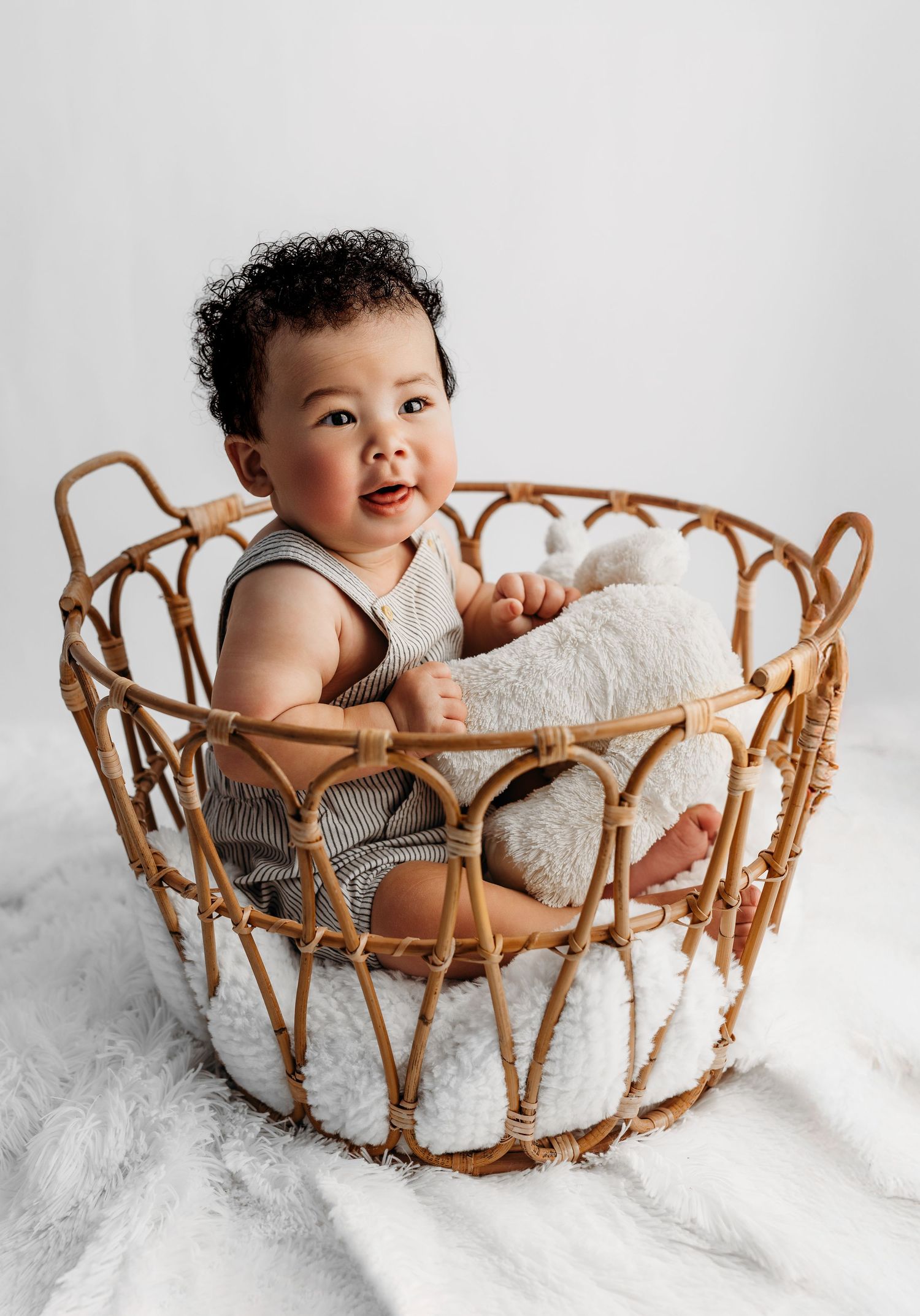 A smiling baby wearing a white dress sits in a vintage-style woven rattan chair against a bright white backdrop.