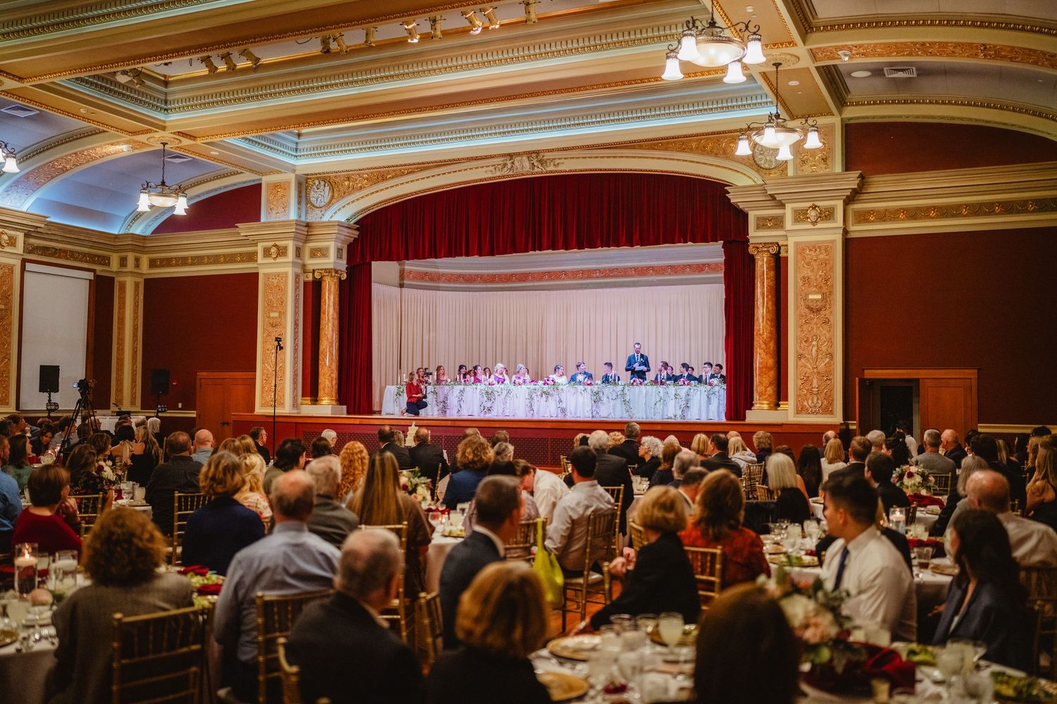A large group of diners seated in an elegant banquet hall with ornate golden decor and chandeliers.
