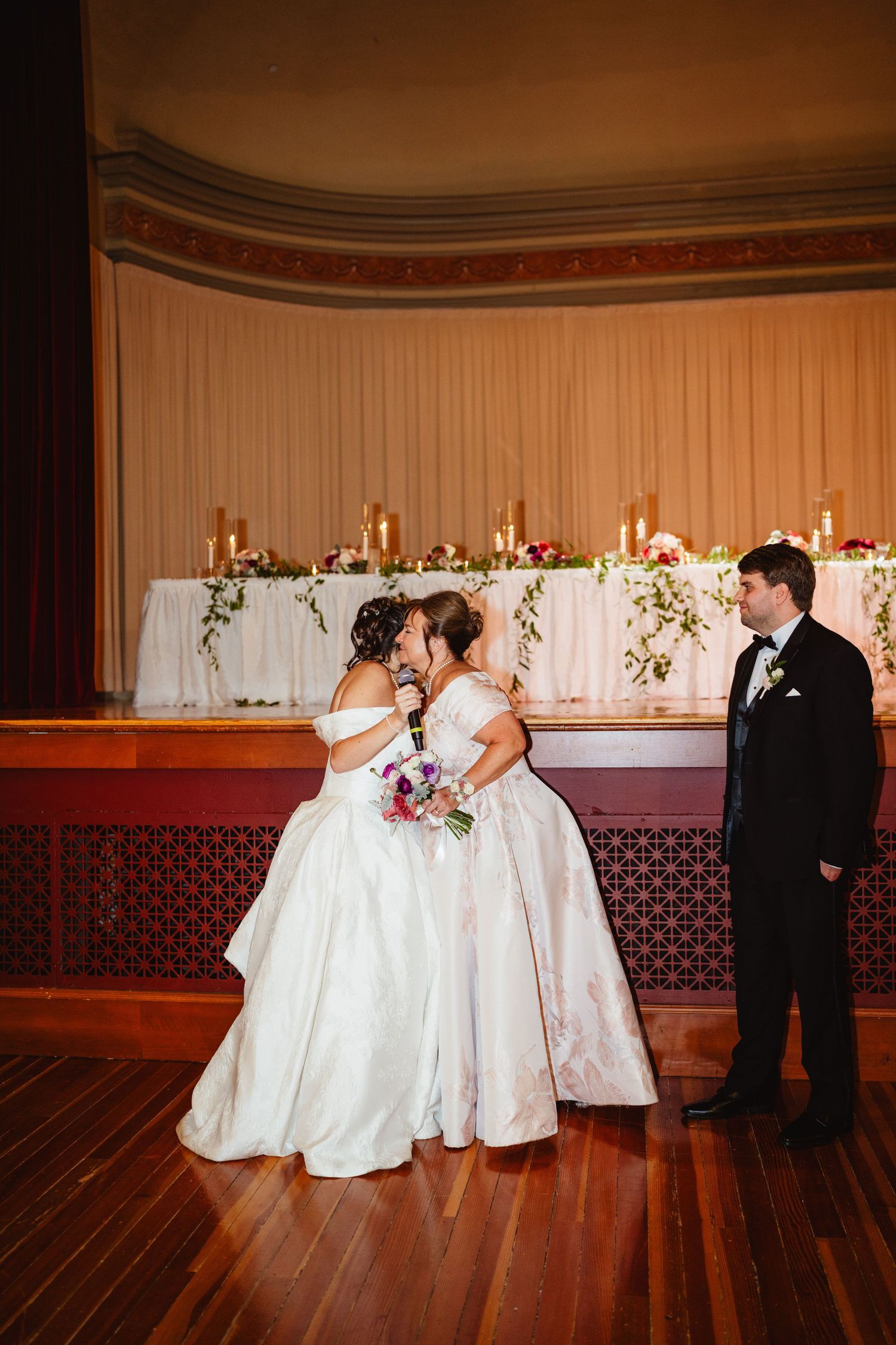 A wedding party dips low in a synchronized dance move on a polished wooden ballroom floor with elegant decor.