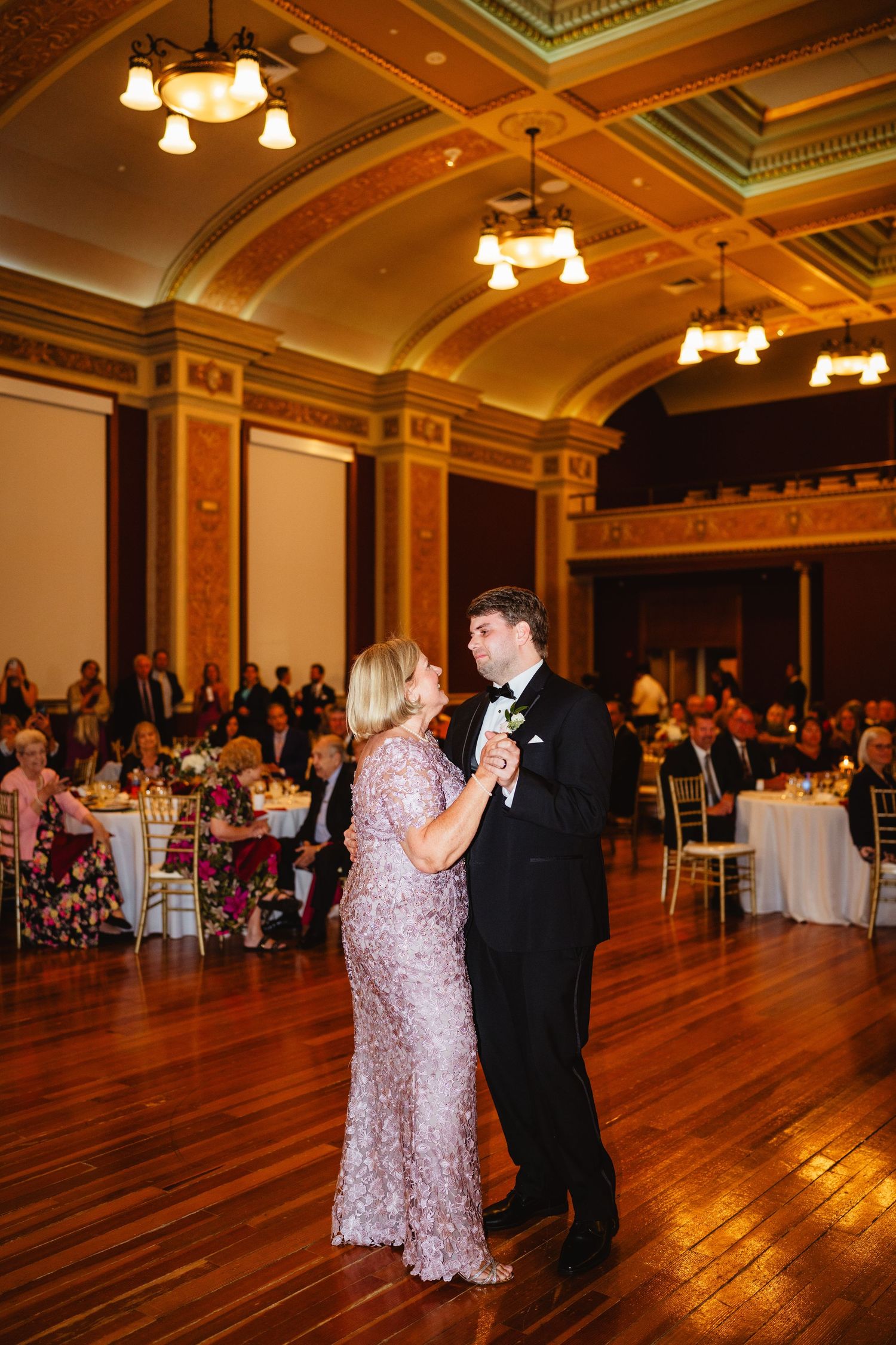 A couple dances gracefully on a polished hardwood floor in an elegant ballroom with ornate moldings and chandeliers.