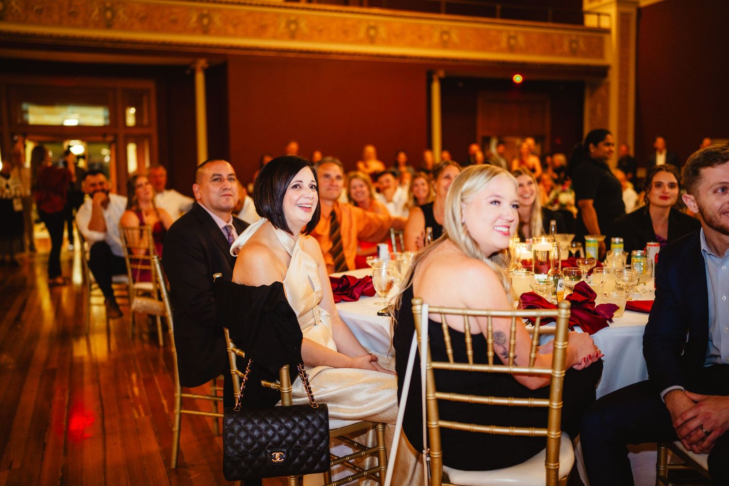 Wedding guests laugh and enjoy an evening reception in a warmly lit ballroom.