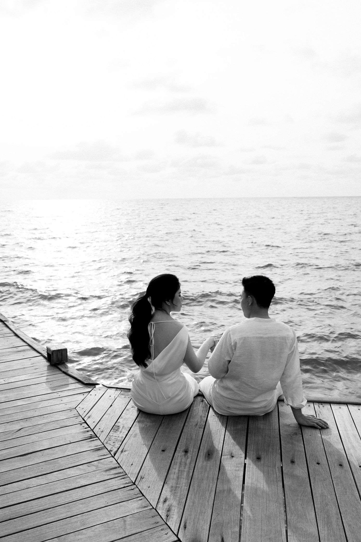Couple sits together on wooden pier gazing at calm ocean in black and white.