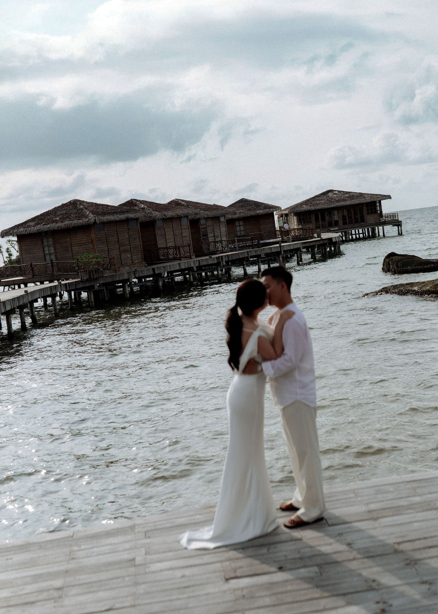 Couple sharing a romantic kiss on a wooden pier with overwater bungalows in the background.