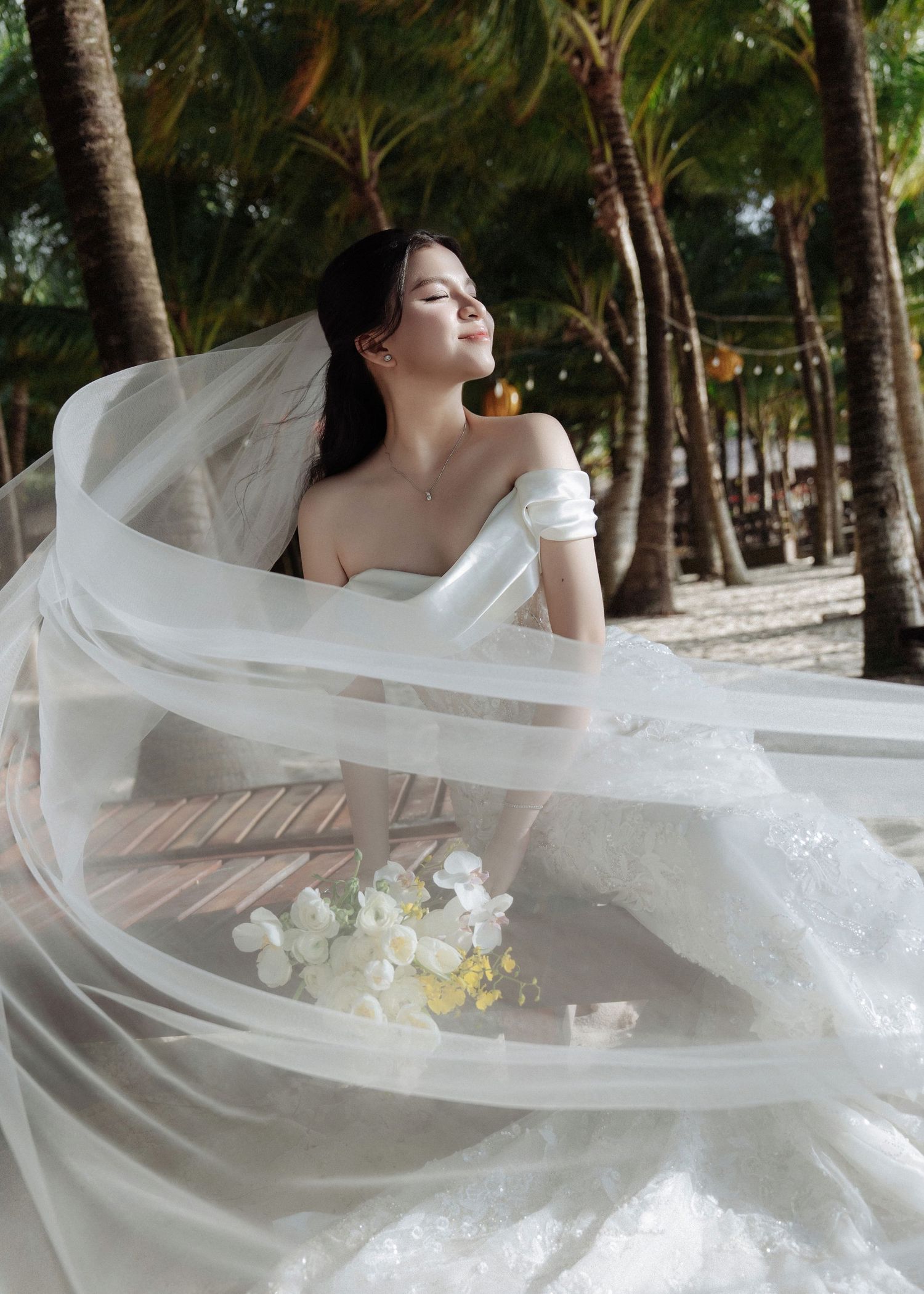 Bride in flowing white wedding gown with veil billowing in the wind among tropical palm trees.