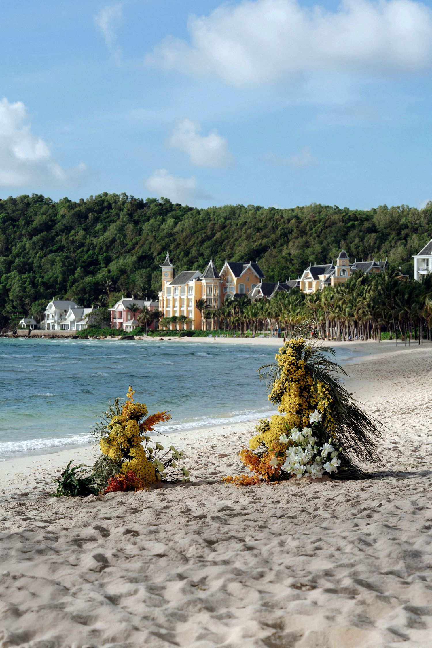 Floral wedding arch arrangements on a sandy beach with colorful resort buildings in the background.