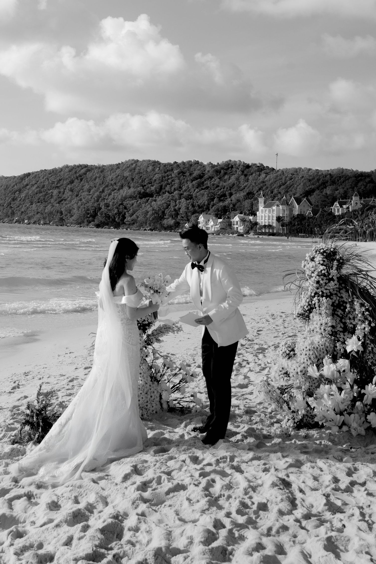 Black and white photo of bride and groom exchanging vows on a beach with floral arch.
