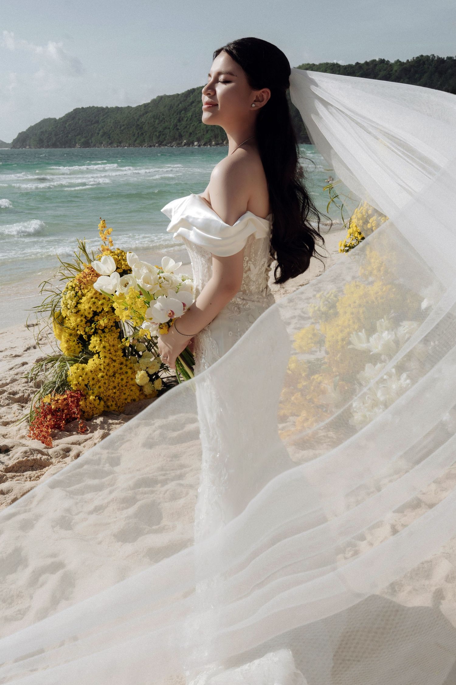 Bride in white off-shoulder gown holding yellow and white bouquet with veil flowing on beach.