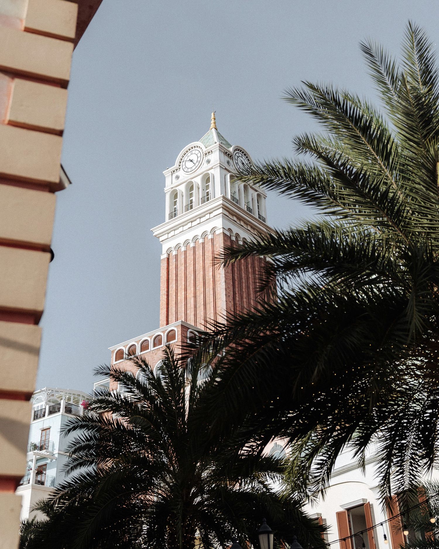 Tall ornate clock tower rising above palm trees against a clear blue sky.