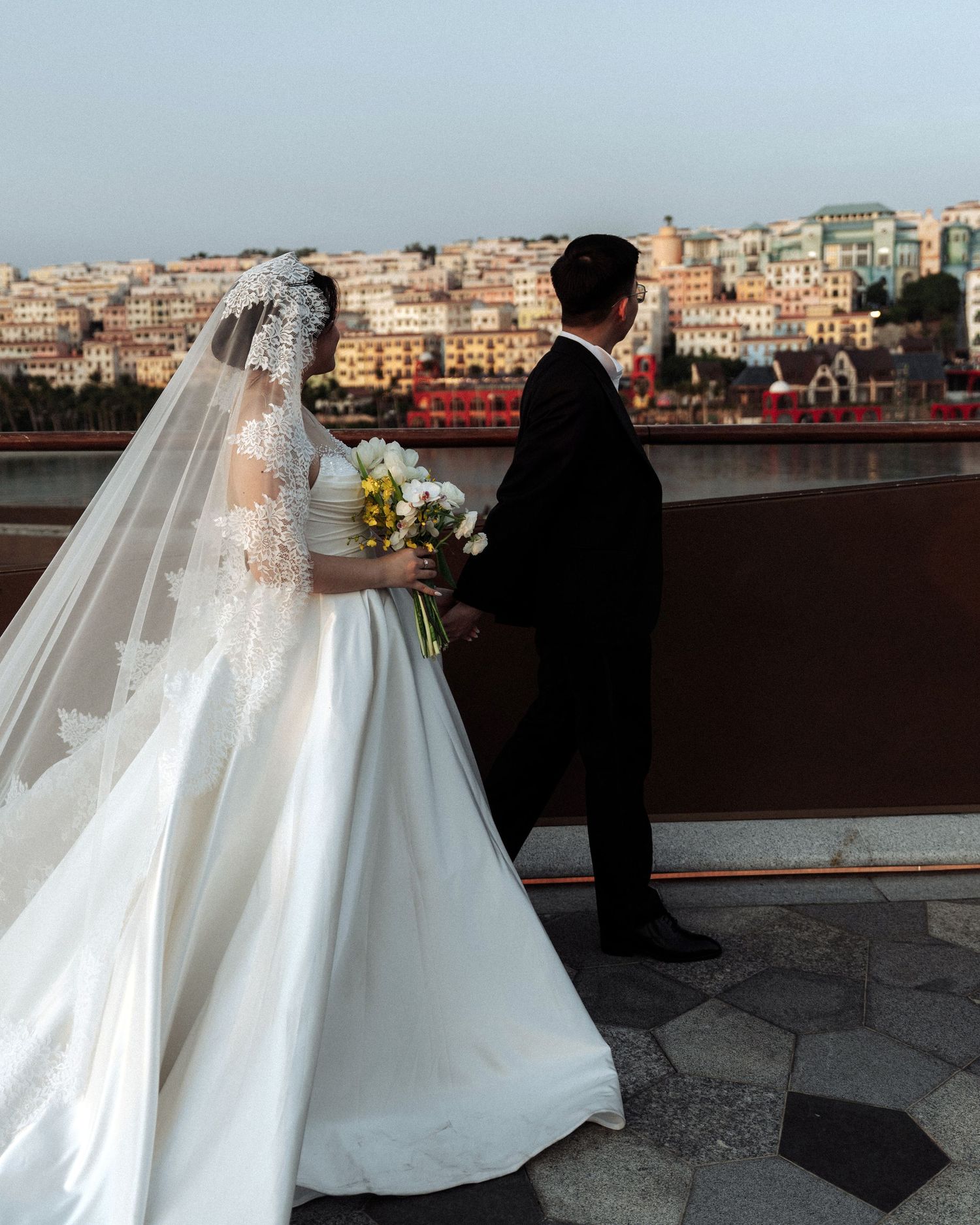 Bride and groom holding hands on a rooftop terrace overlooking a colorful coastal cityscape.