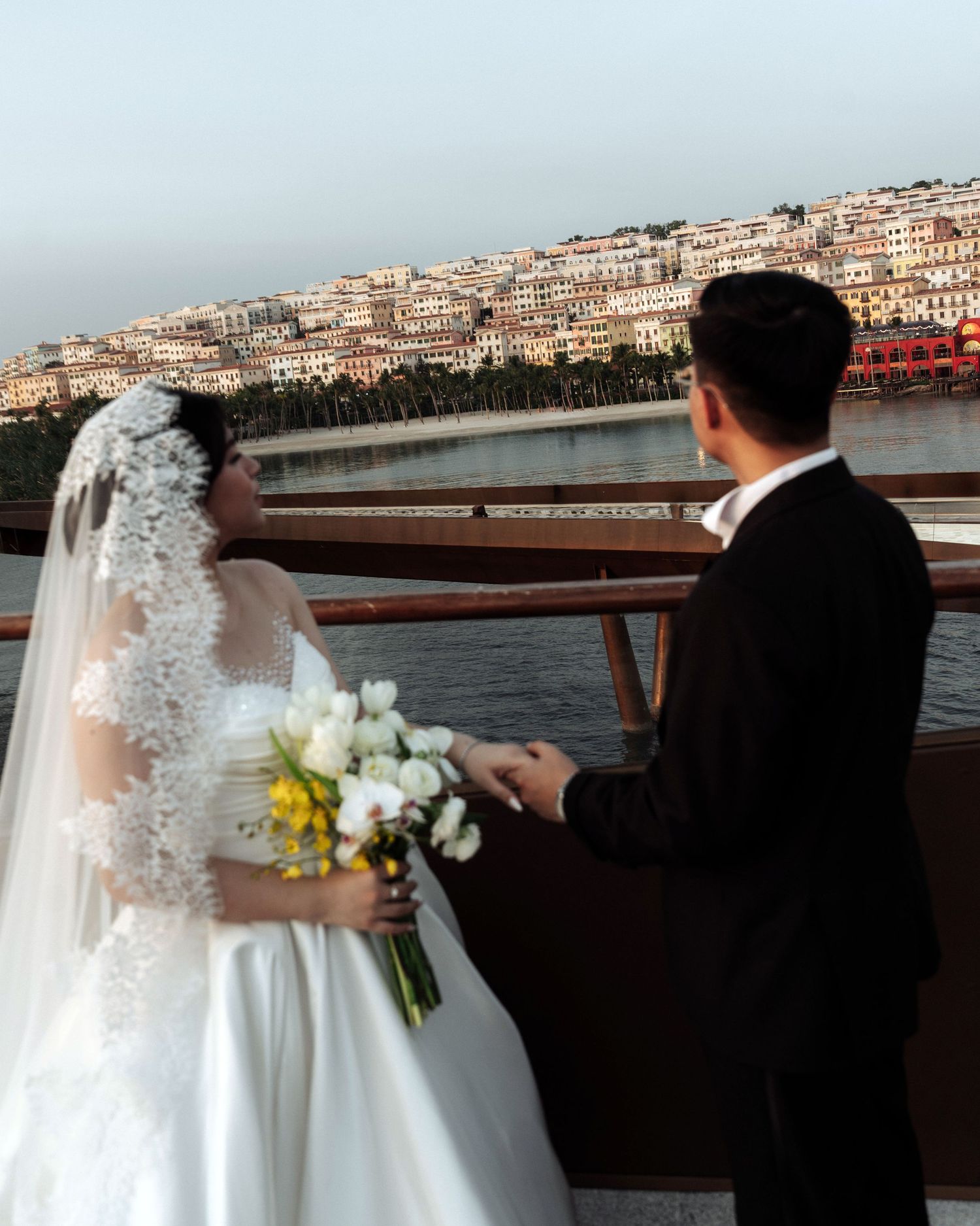 Bride holding white bouquet facing groom on a boat deck with scenic waterfront city view.