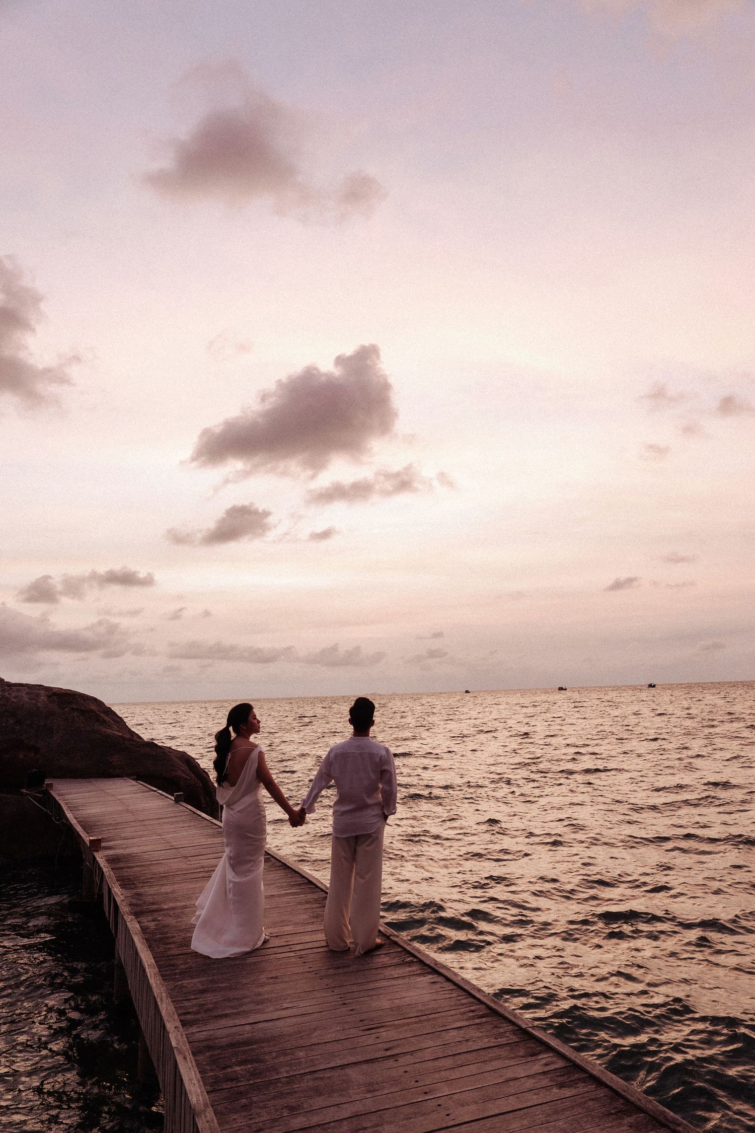 Couple holding hands on a wooden pier at sunset over the ocean.