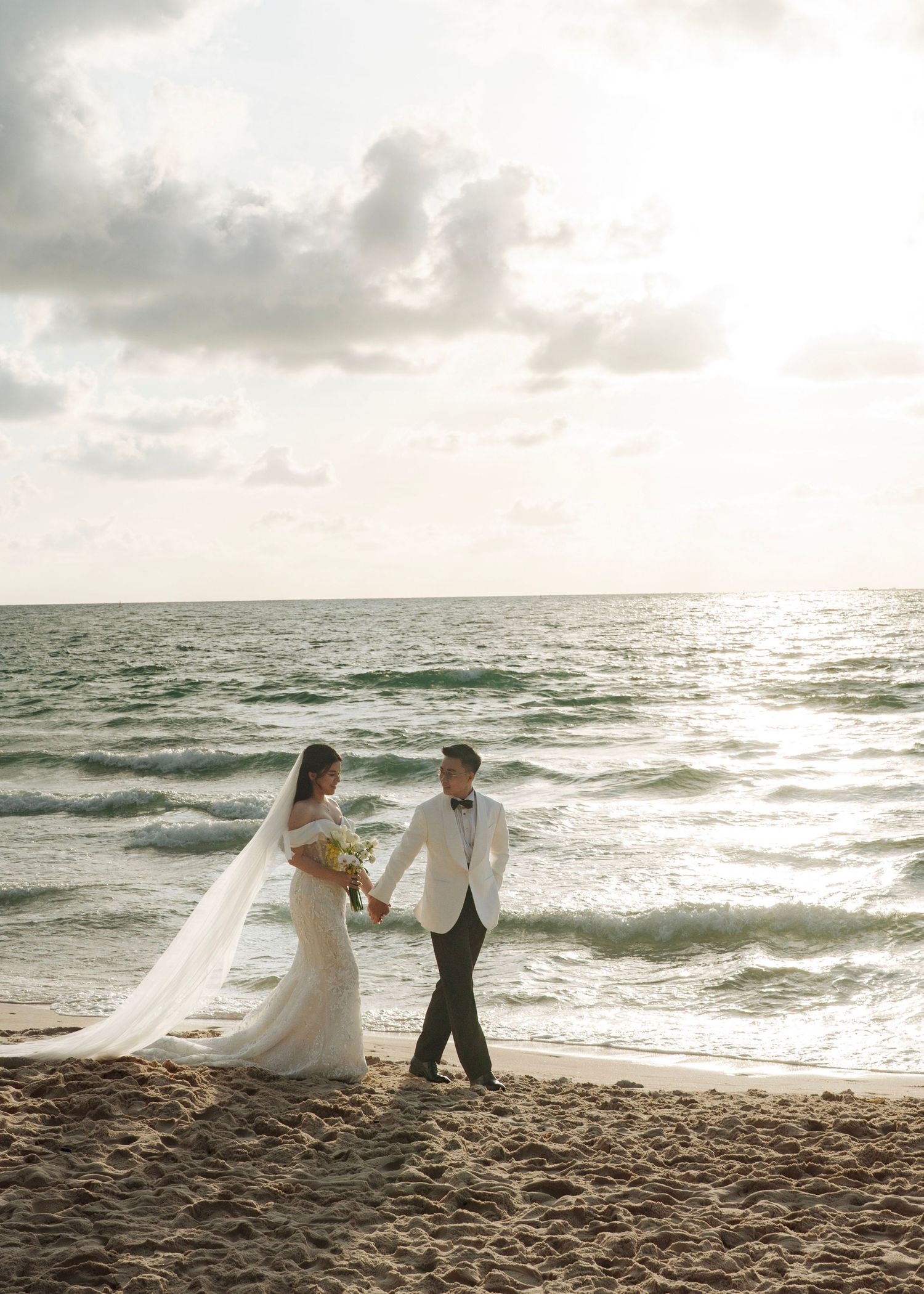 Bride and groom walk hand in hand along a sunlit beach.