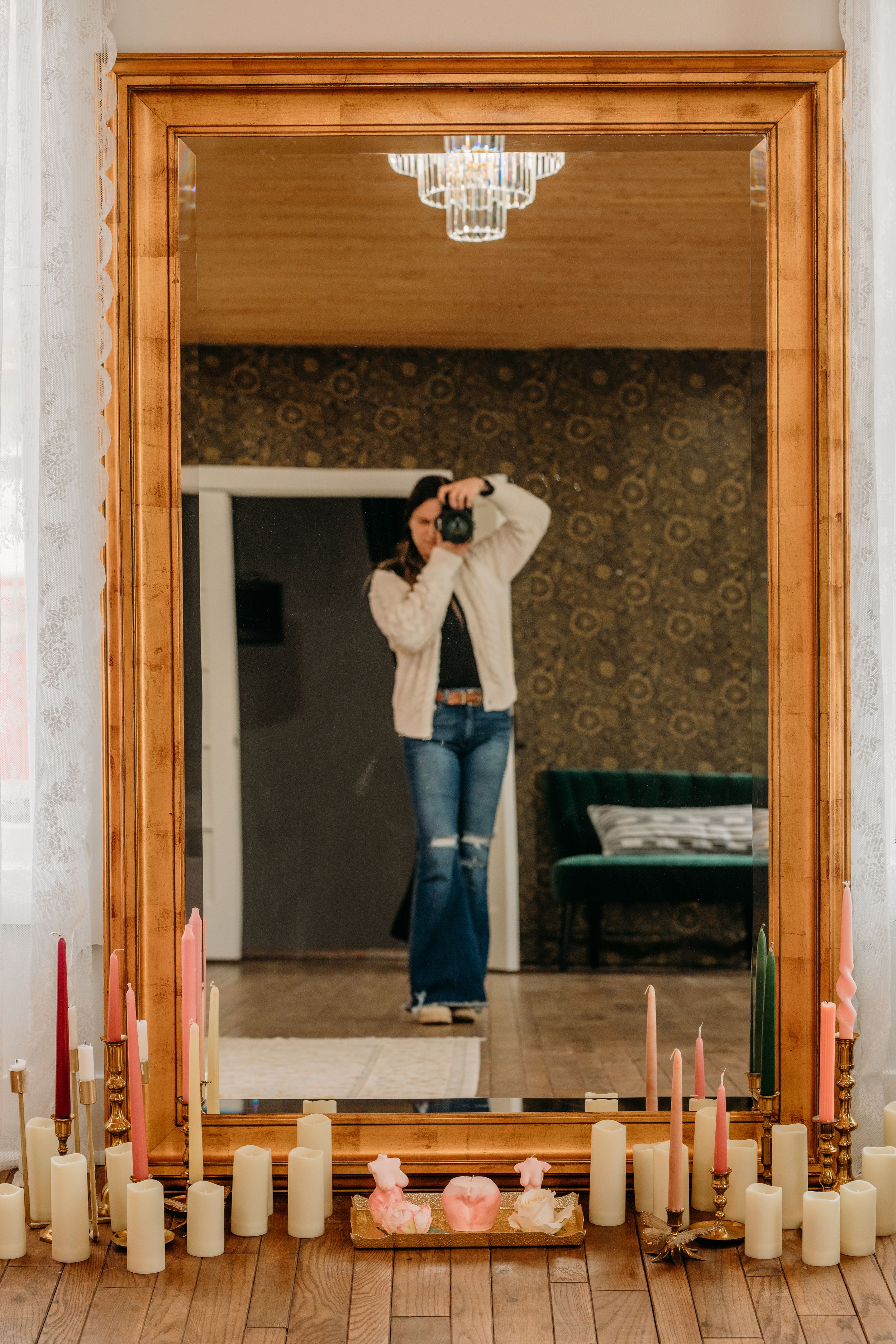 Photographer taking a mirror selfie surrounded by candles in a decorated room with a large gold-framed mirror.