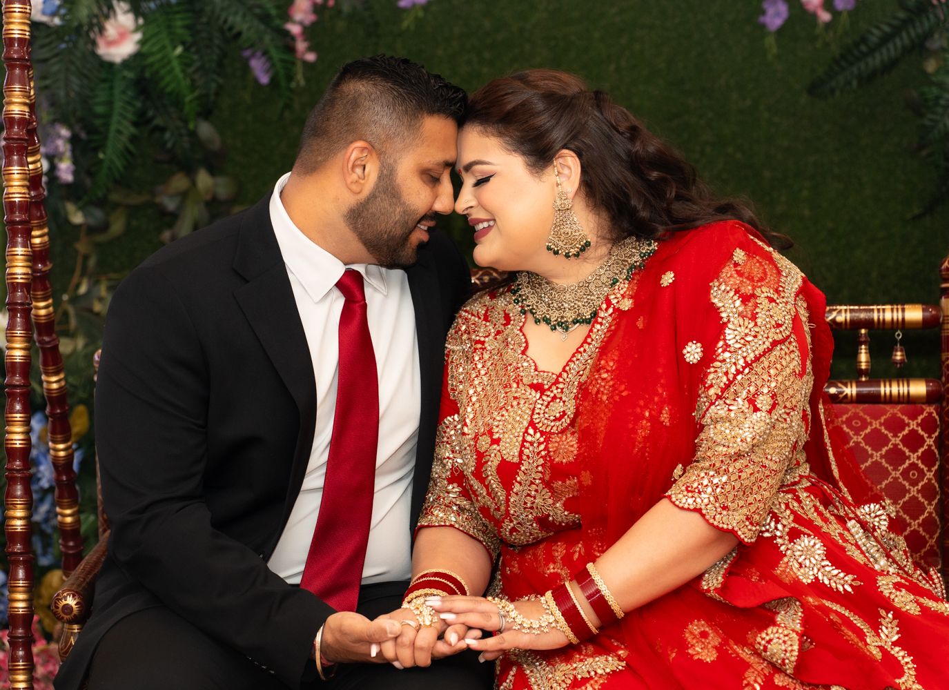 A romantic couple in traditional South Asian wedding attire share intimate moments against a green floral backdrop.