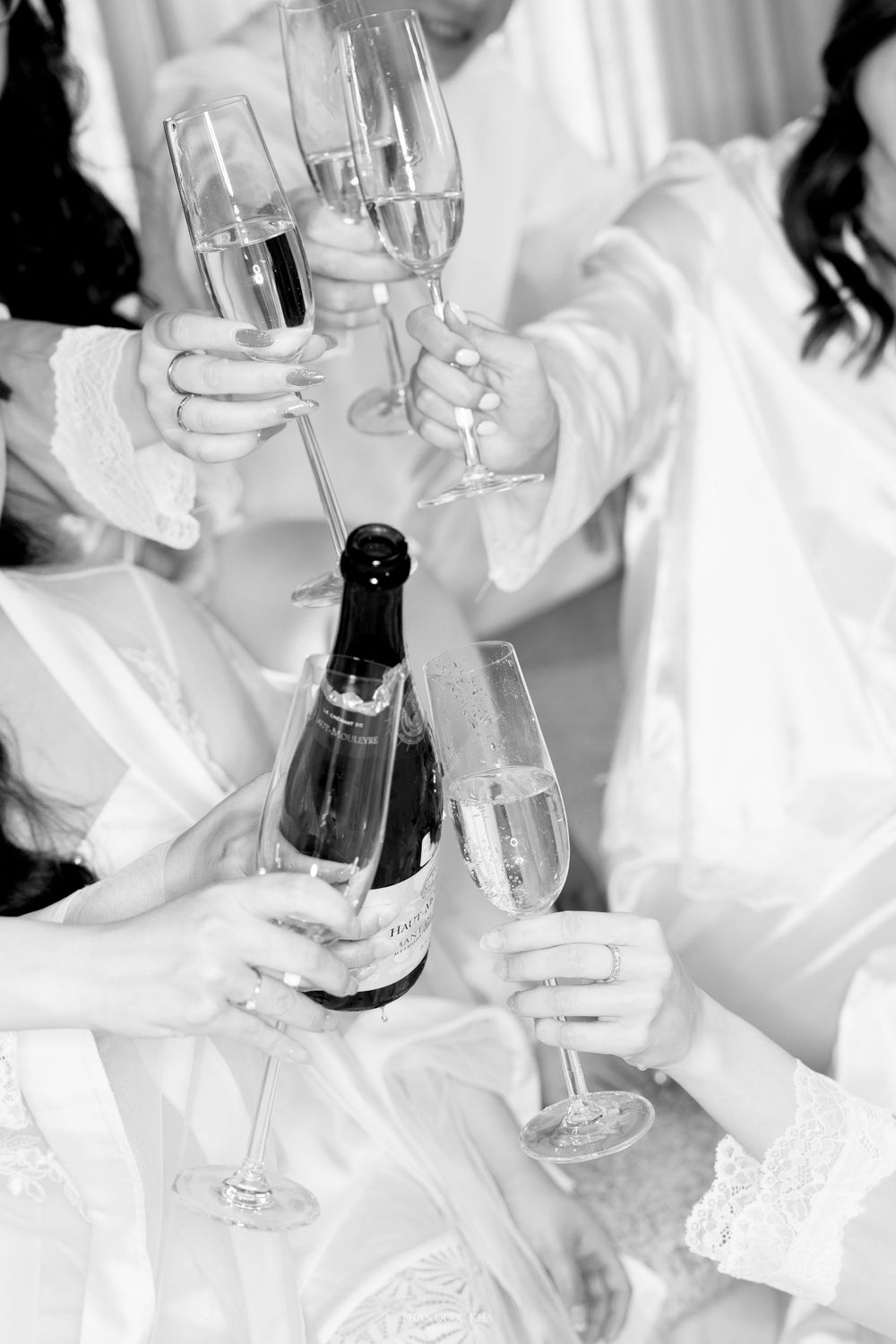 Black and white close-up of wedding party clinking champagne glasses in white robes before ceremony.