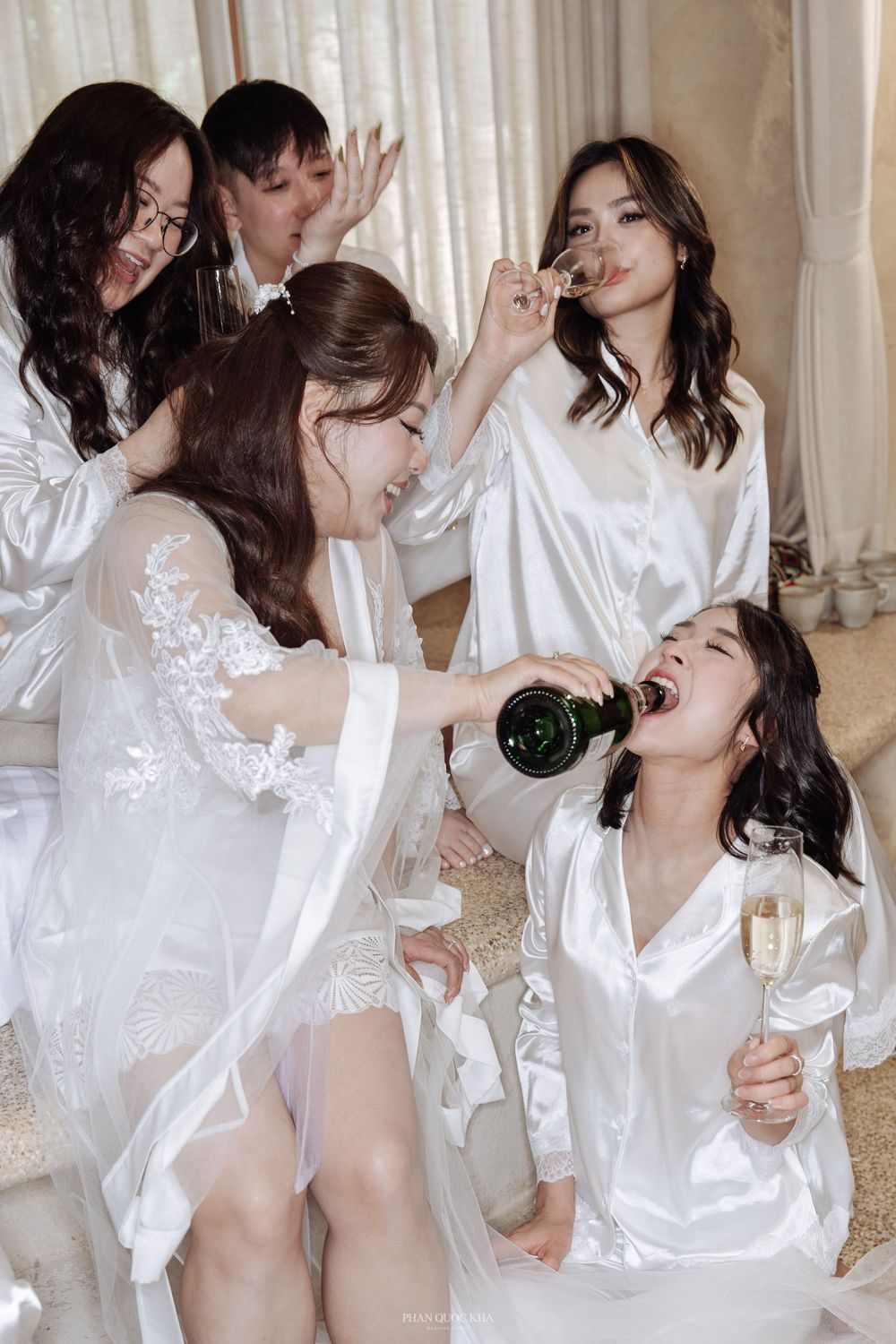 Bridesmaids in white satin robes laugh as champagne is poured during fun bridal morning preparations.