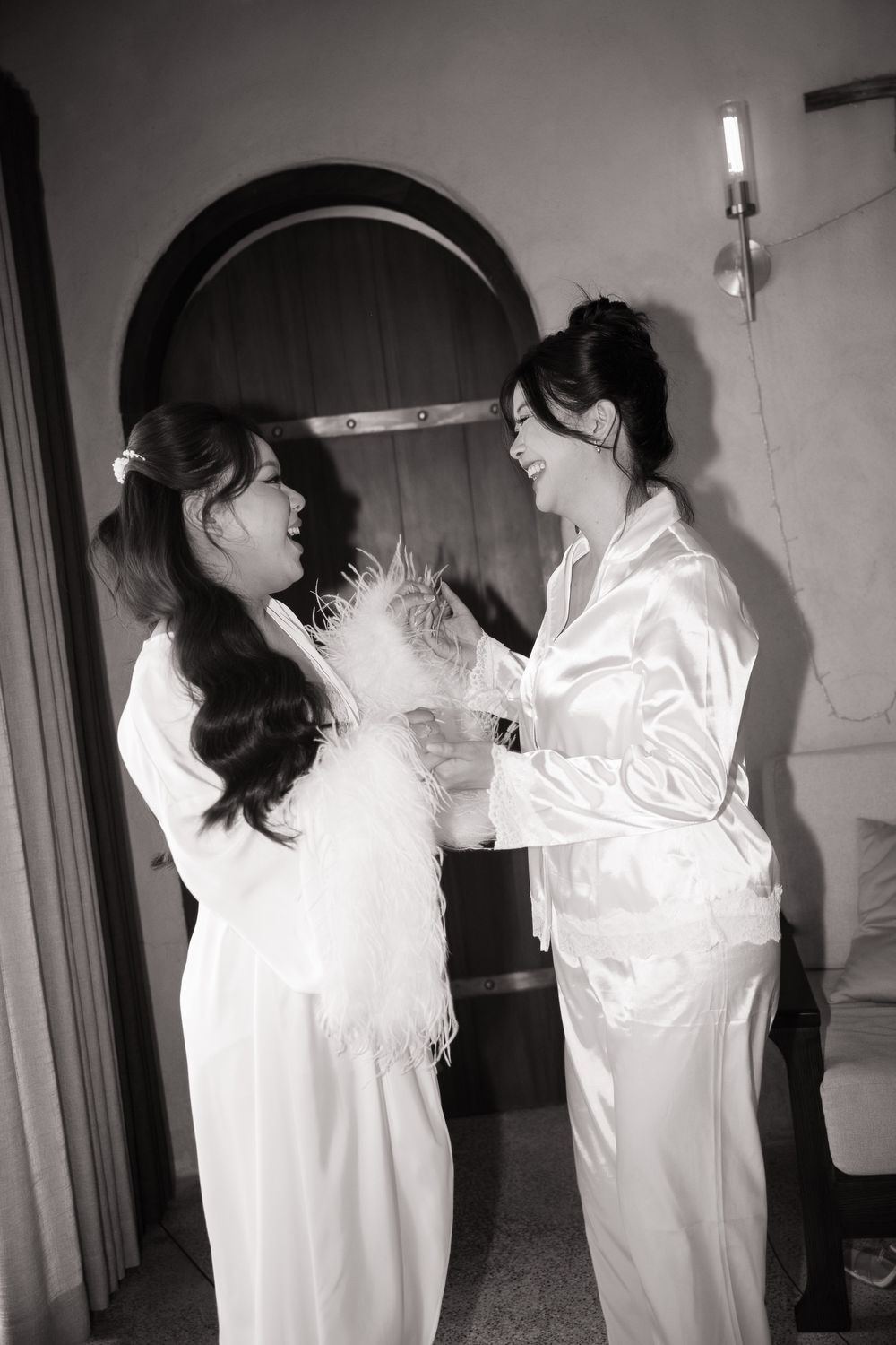Black and white photo of two women in white robes laughing and sharing a joyful bridal moment.