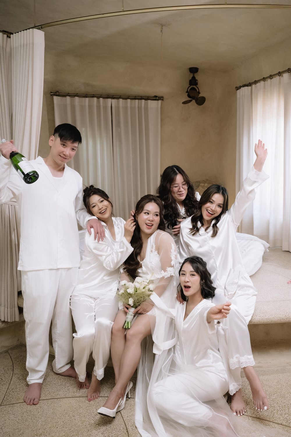 Bride and bridesmaids in white satin robes celebrate with champagne and bouquet in a bright hotel room.