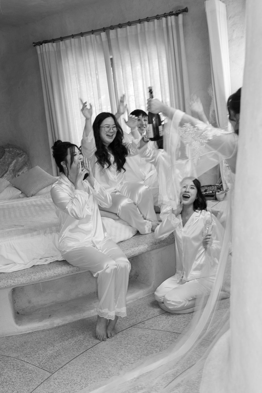 Black and white photo of bridesmaids in white satin pajamas celebrating with champagne on a bed.