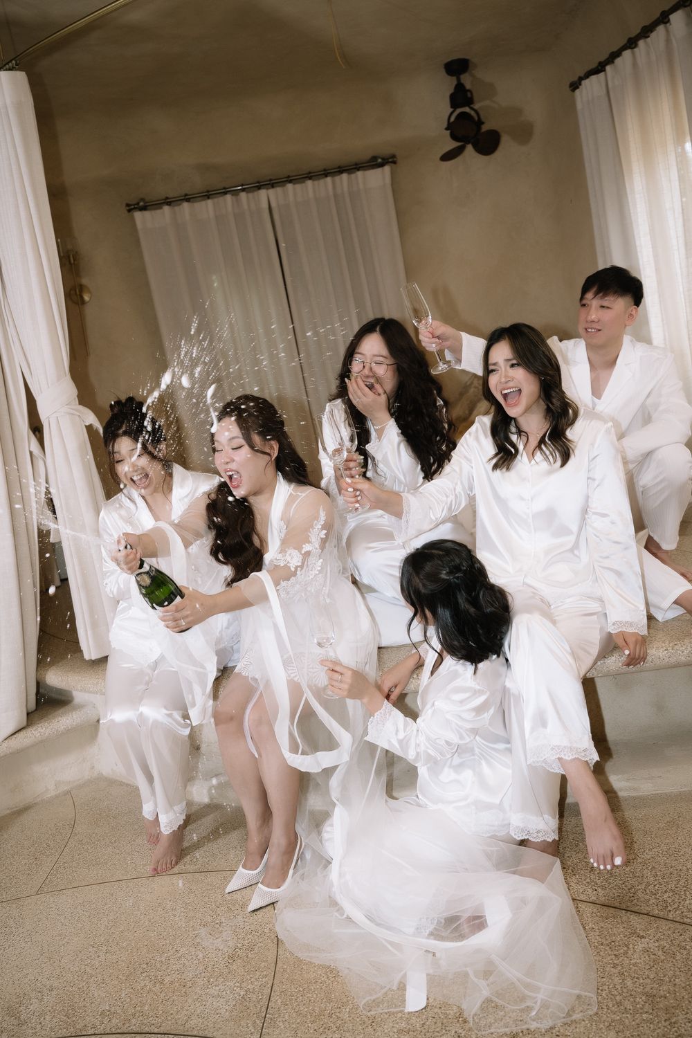 Excited bridal party in white robes reacts to champagne bottle popping during wedding preparations.
