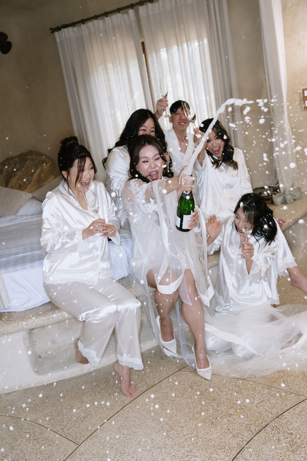 Bride and bridesmaids in white satin robes celebrate with champagne spray during bridal preparations.