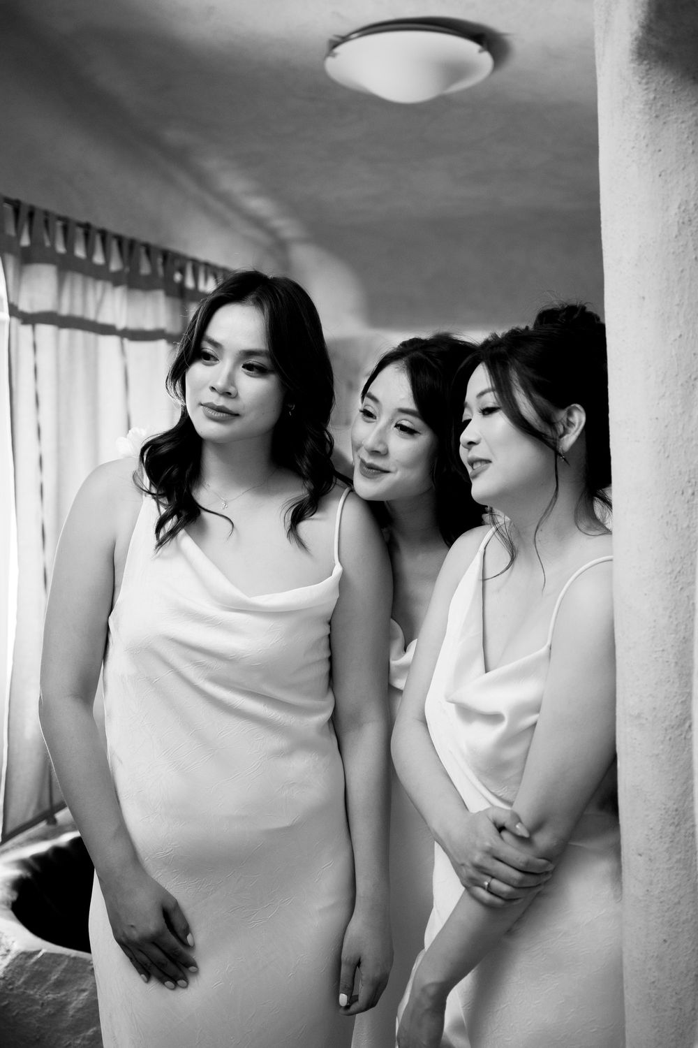 Black and white portrait of three bridesmaids in white slip dresses posing together indoors.