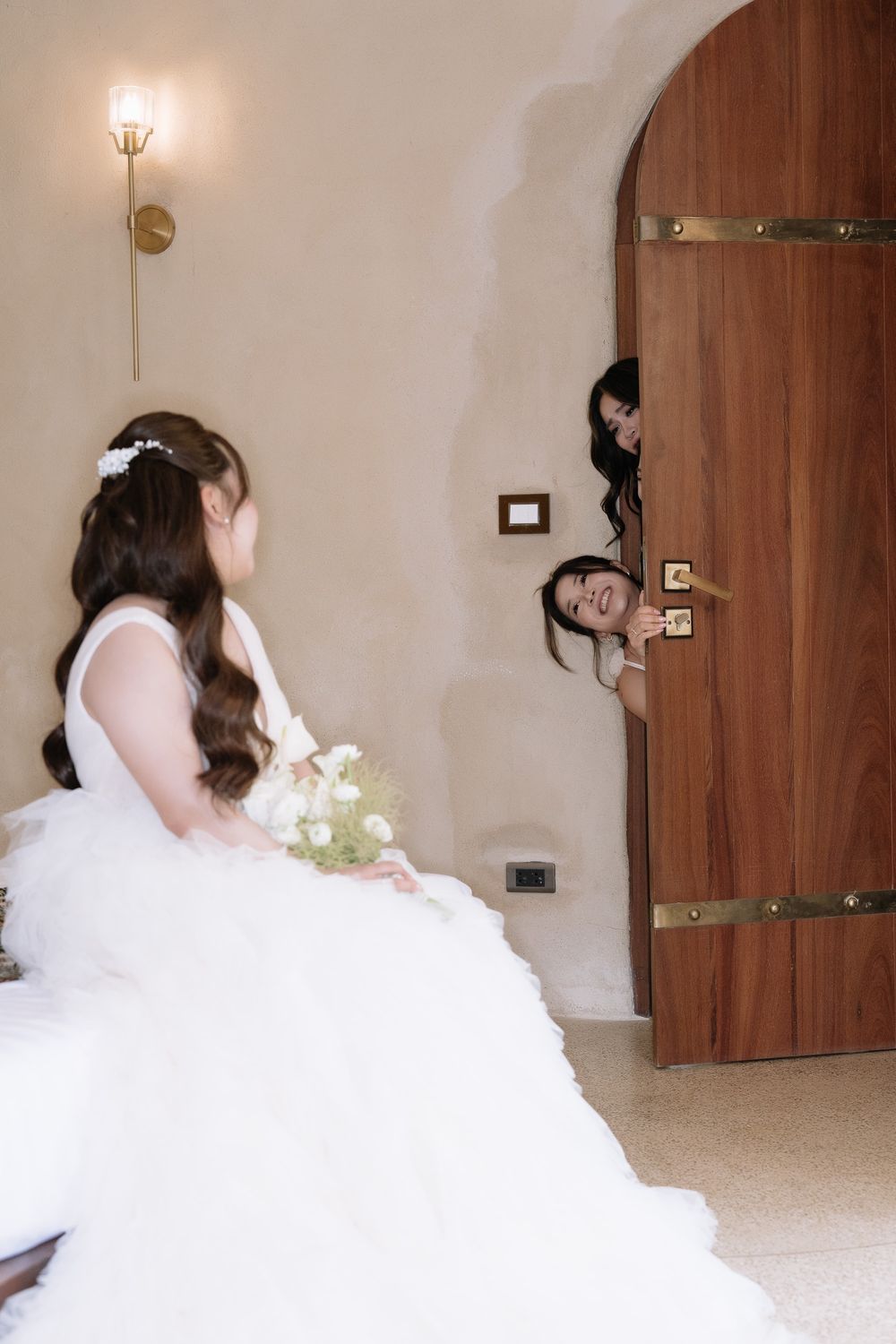 Bride in full white gown holds bouquet as bridesmaids peek through arched wooden door smiling.
