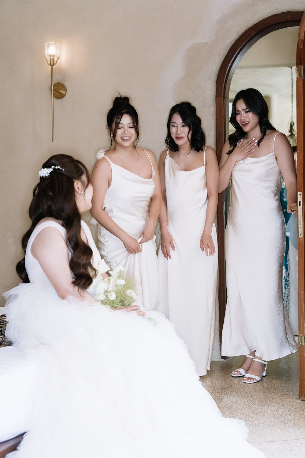 Bride in white gown sits as three bridesmaids in white slip dresses admire her in doorway.