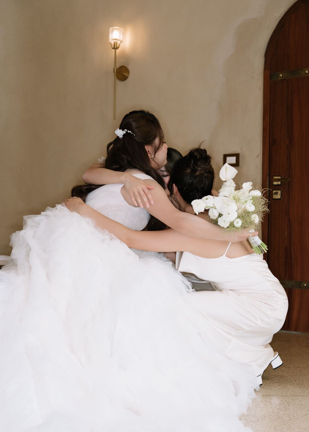 Bride in a white tulle gown sharing a heartfelt hug with bridesmaids while holding a white floral bouquet.