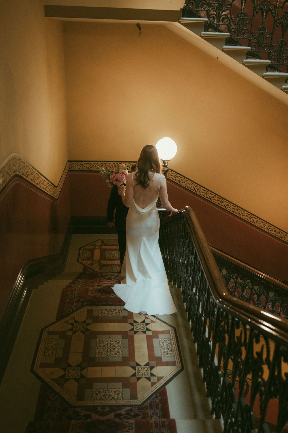 Bride in white gown holding pink bouquet descends ornate staircase with iron railings and decorative tile floor.