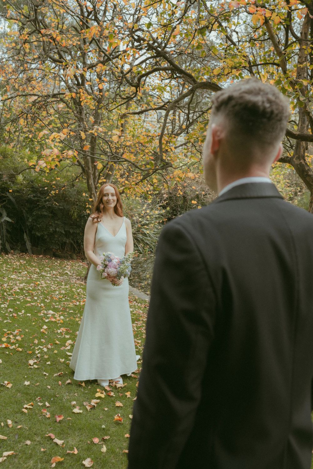Bride in white gown holds bouquet facing groom in dark suit during autumn outdoor wedding ceremony.