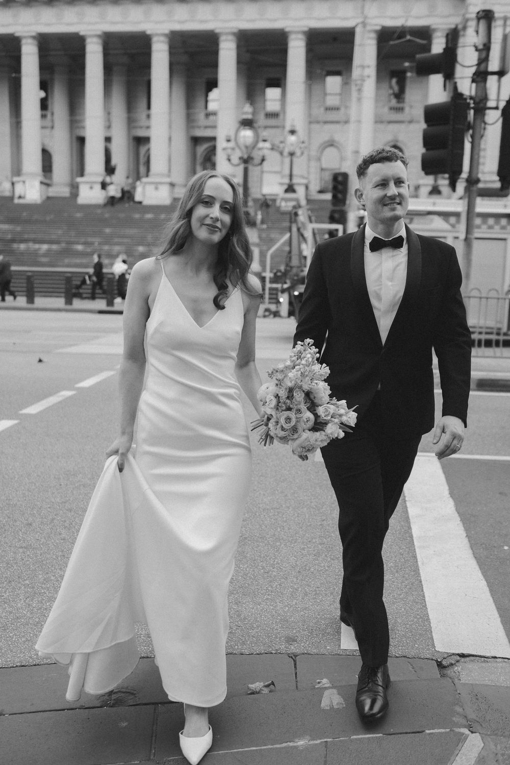 Black and white photo of a smiling bride in white gown and groom in tuxedo walking together outside a grand building.