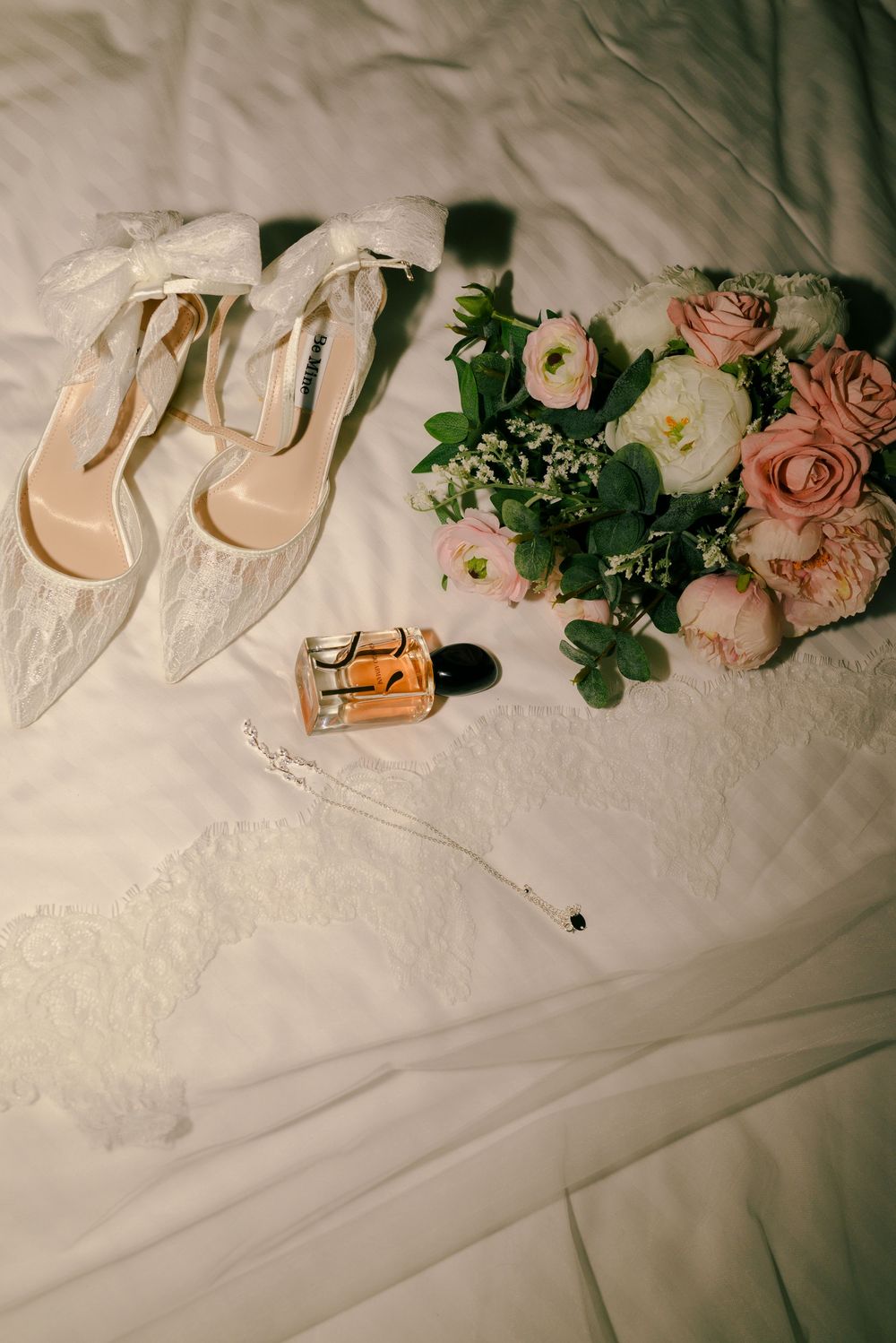 Bridal flat lay with lace heels, pink and white bouquet, perfume bottle, and white lace wedding dress on bed.