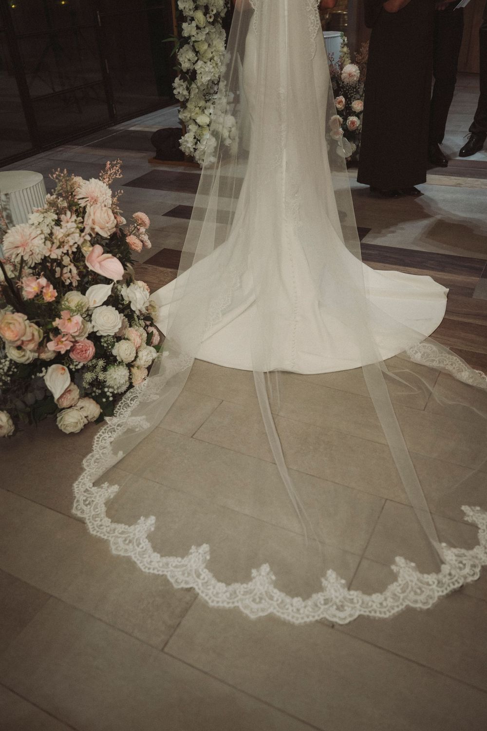 Elegant bride in white gown with lace-trimmed cathedral veil trailing on marble floor beside lush pink and white floral arrangements.