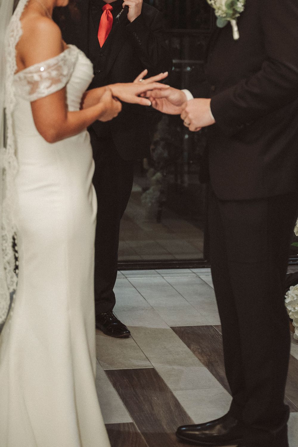 Bride and groom exchanging rings during wedding ceremony, bride in white off-shoulder gown with veil.