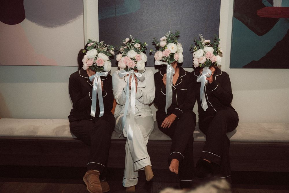 Four people in dark pajamas sit together holding large floral bouquets covering their faces, in an artistic posed photo.