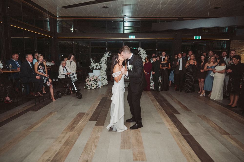 Bride and groom share their first dance on a wooden dance floor surrounded by wedding guests at an elegant reception venue.
