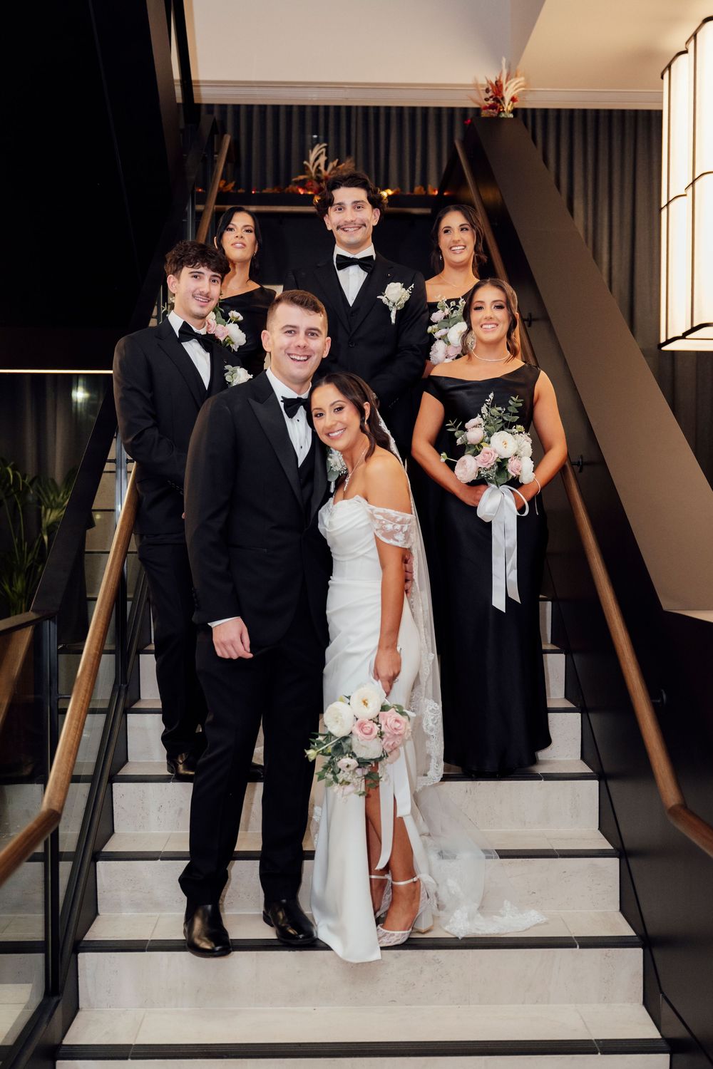 Wedding party poses on elegant staircase, bride in white gown with bouquet, groom in tuxedo, bridesmaids in navy dresses.