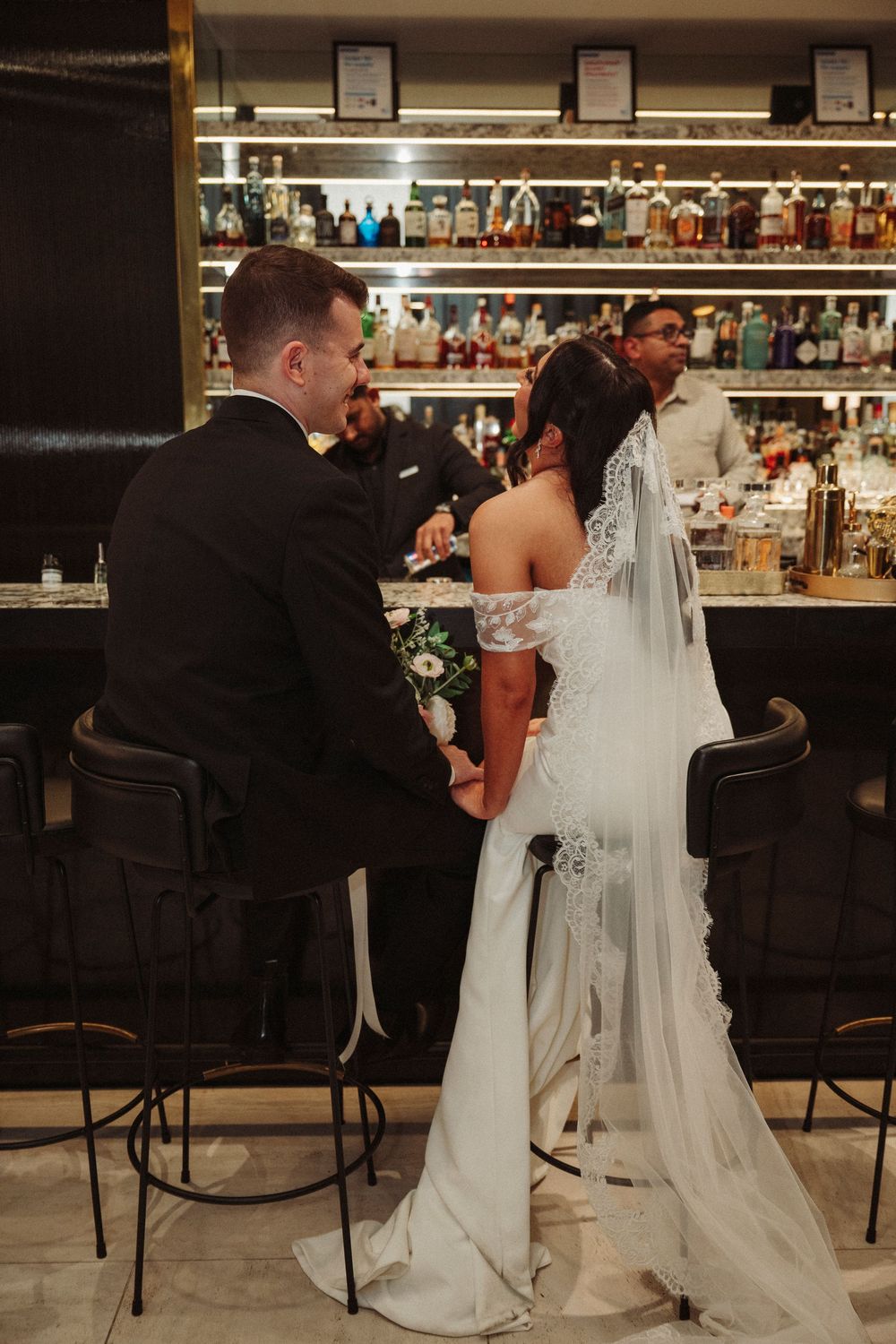 Bride and groom sit together at an elegant bar, holding hands in wedding attire, with a bartender serving in the background.
