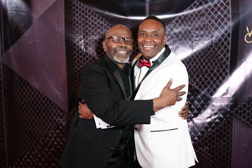 Two people in formal attire embracing and smiling warmly at an event in front of a chain-link pattern backdrop.