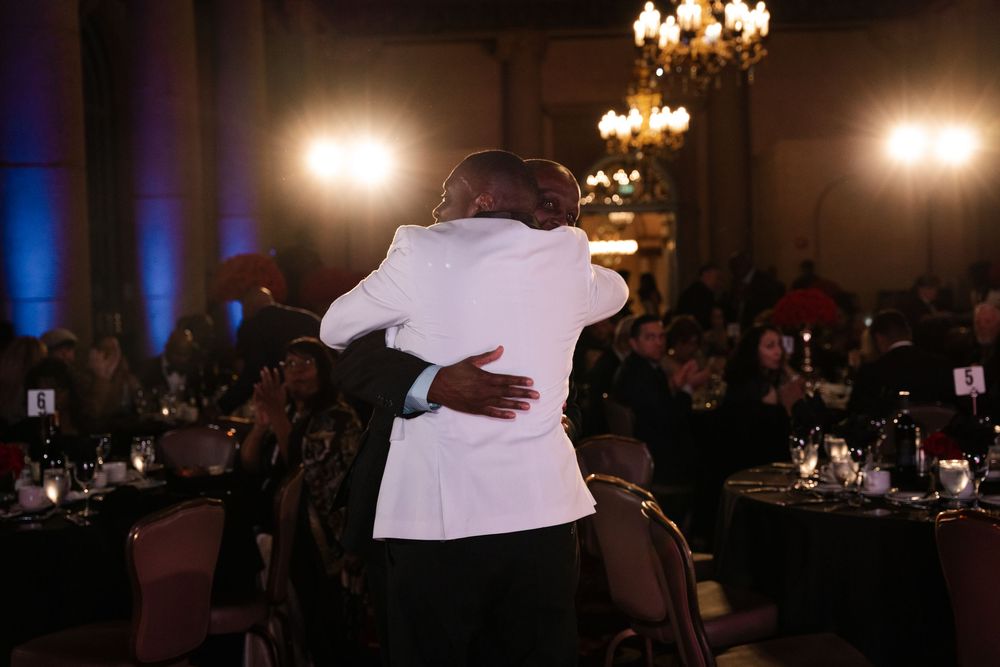 People dance together in a dimly lit ballroom at a formal event with elegant chandeliers overhead.