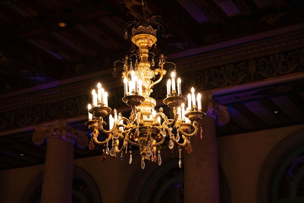 An elegant crystal and gold chandelier with multiple lit candles illuminates a dark ornate ceiling.
